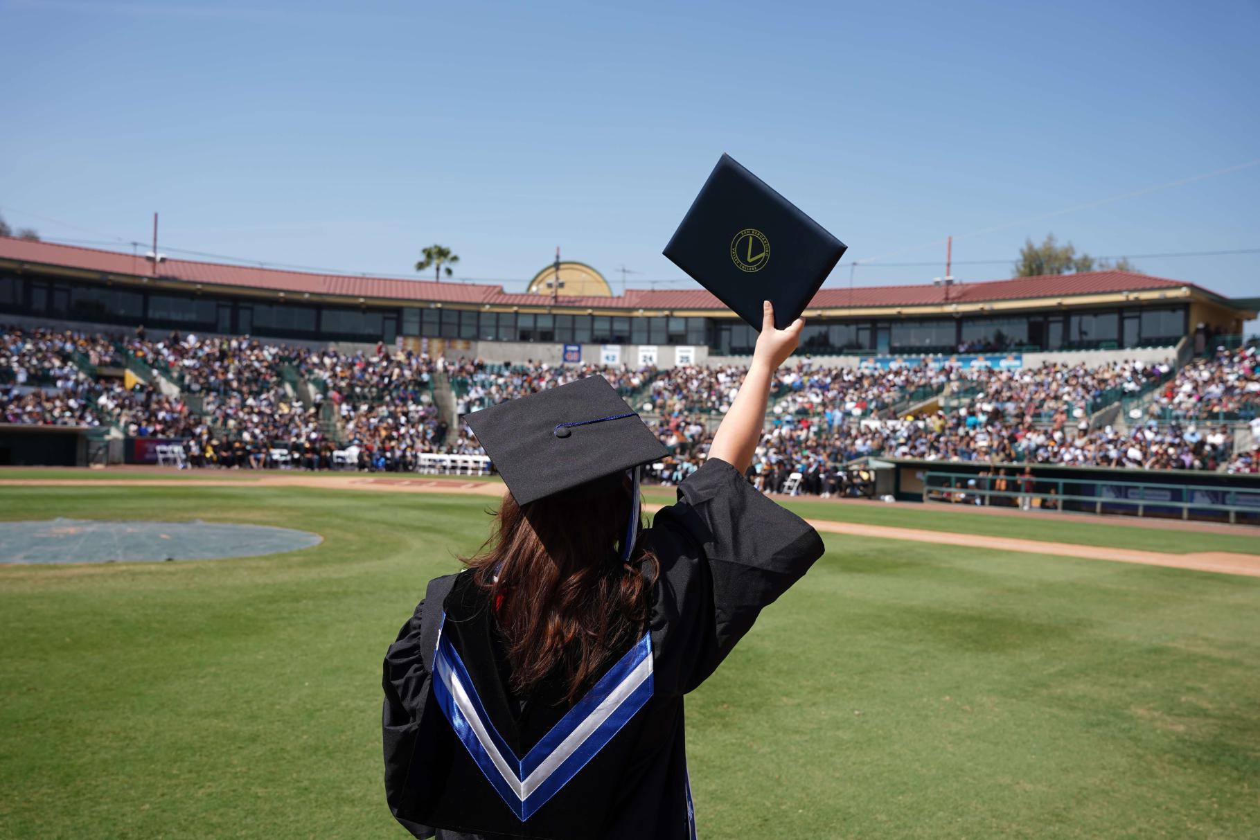 A graduate holding a diploma cover stands with her back to the stadium while raising it high in the air above her head.