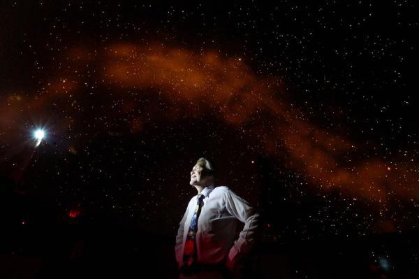 A photo of Chris Clarke standing in the planetarium while the stars are projected on and above him.