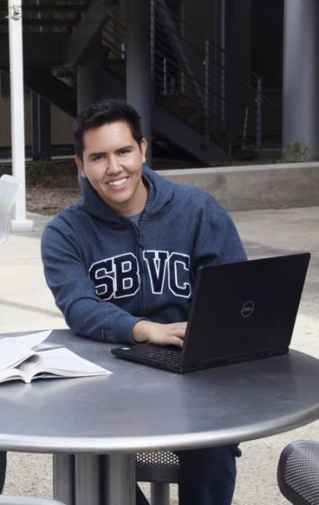A photo of Oscar Luna in a SBVC hoodie on a laptop outside the Campus Center building.