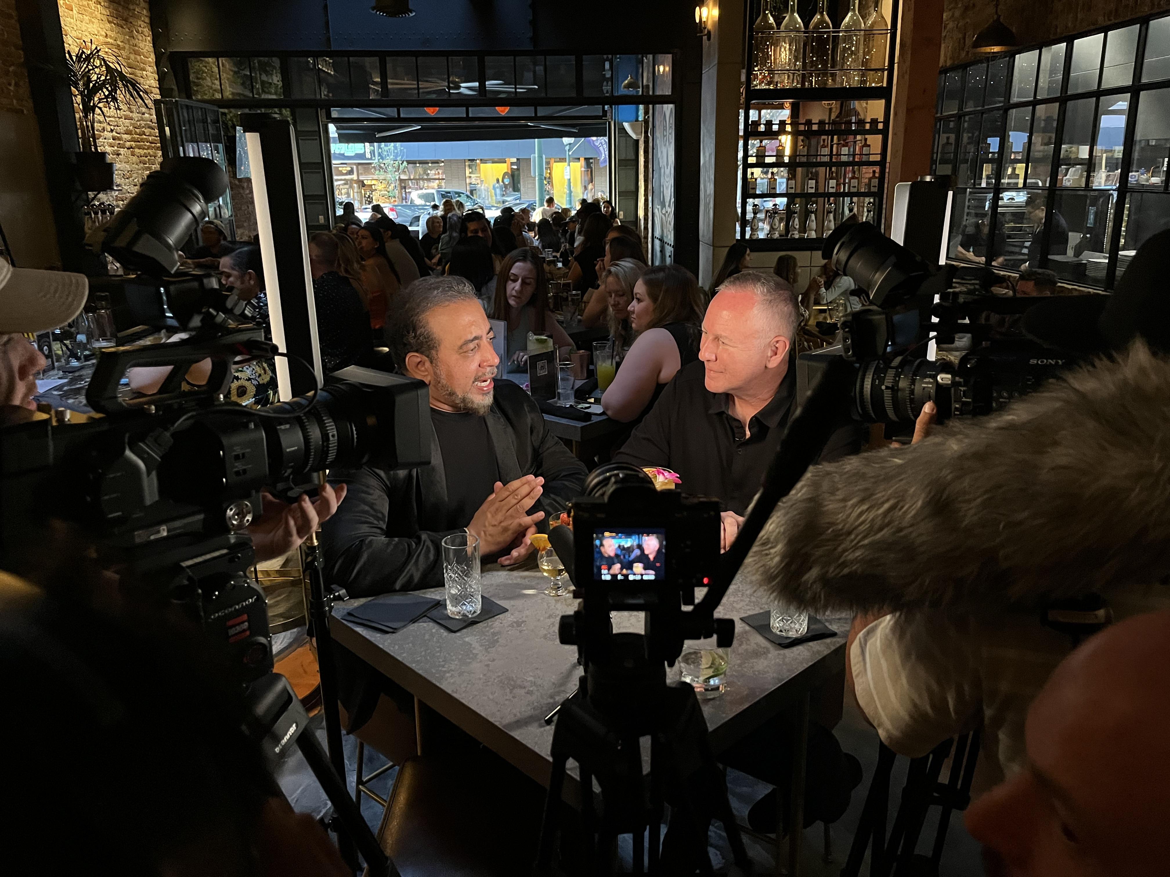 Two men being filmed speaking in a bar.