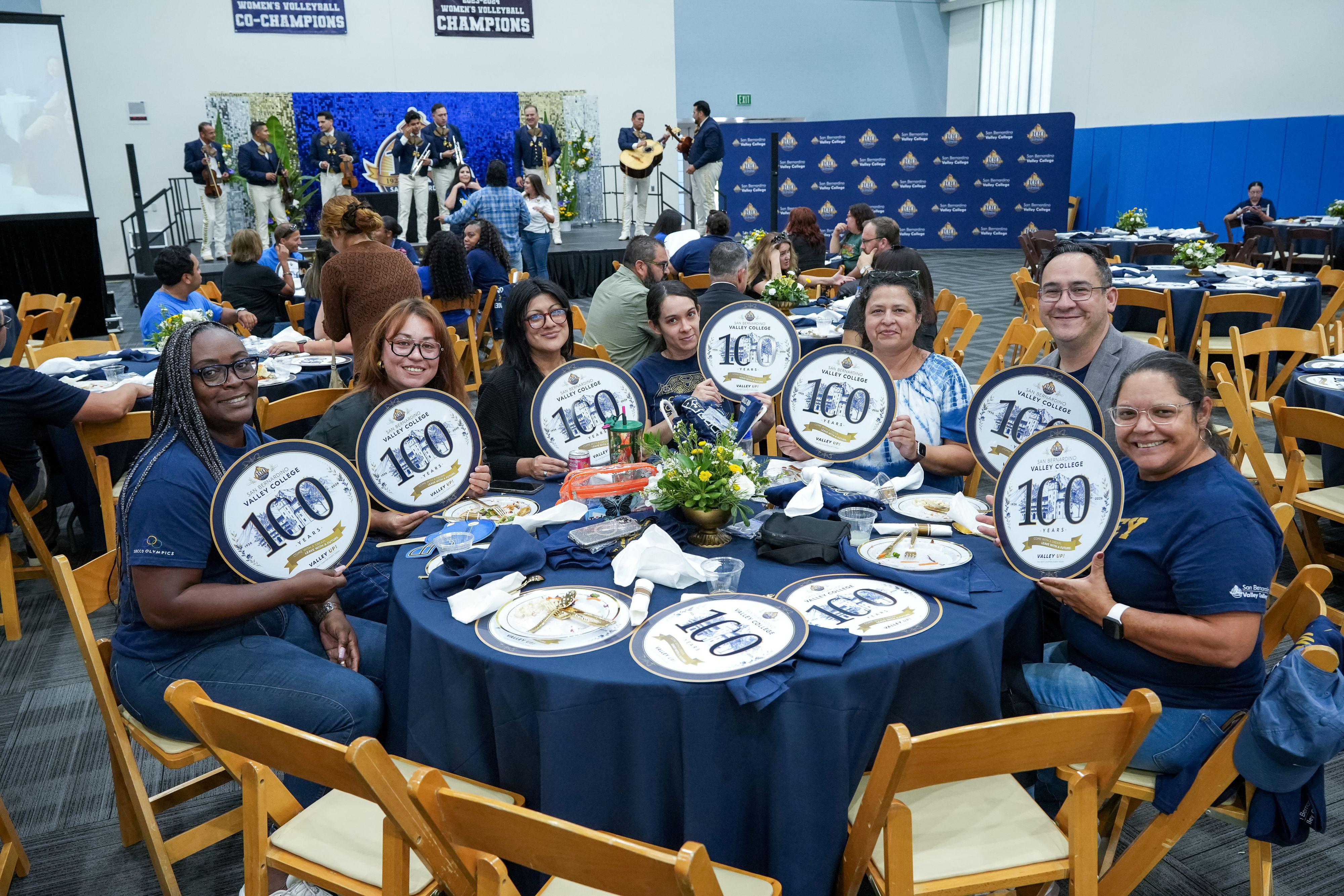 A group of people sit around a table holding SBVC centennial chargers