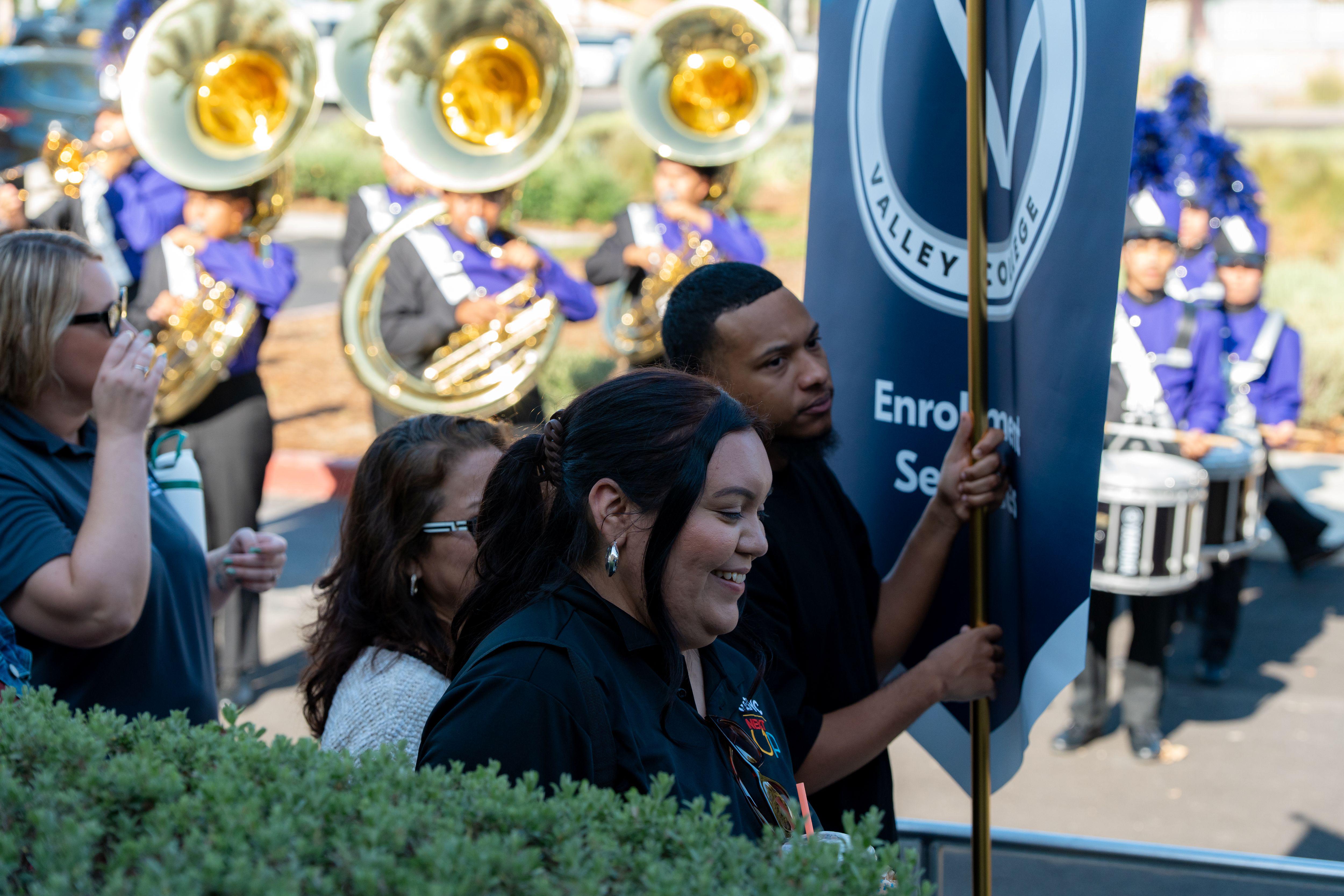 A few people walk with a marching band behind them, a man holds a blue banner