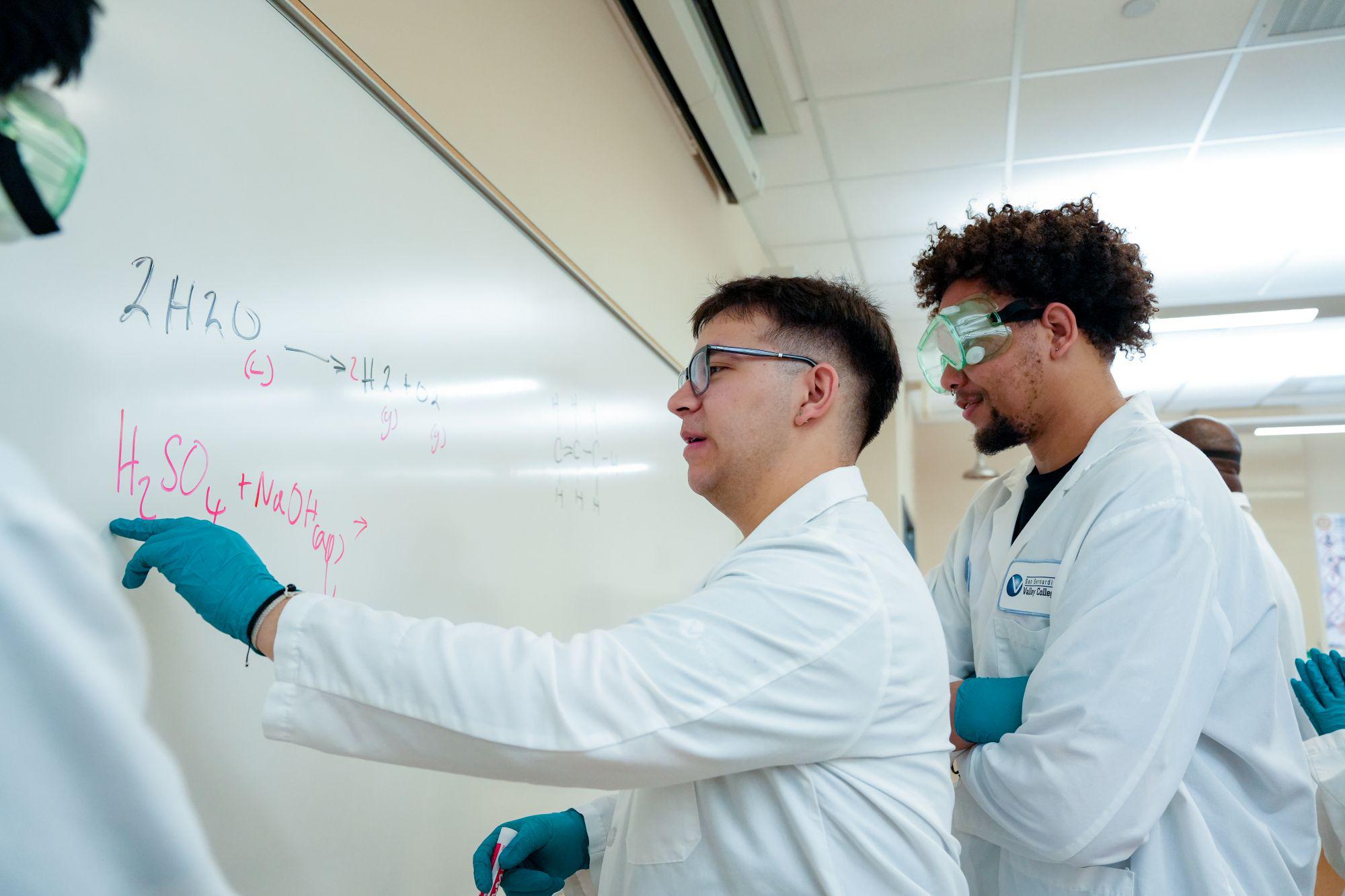 Two male students write chemical elements on a whiteboard