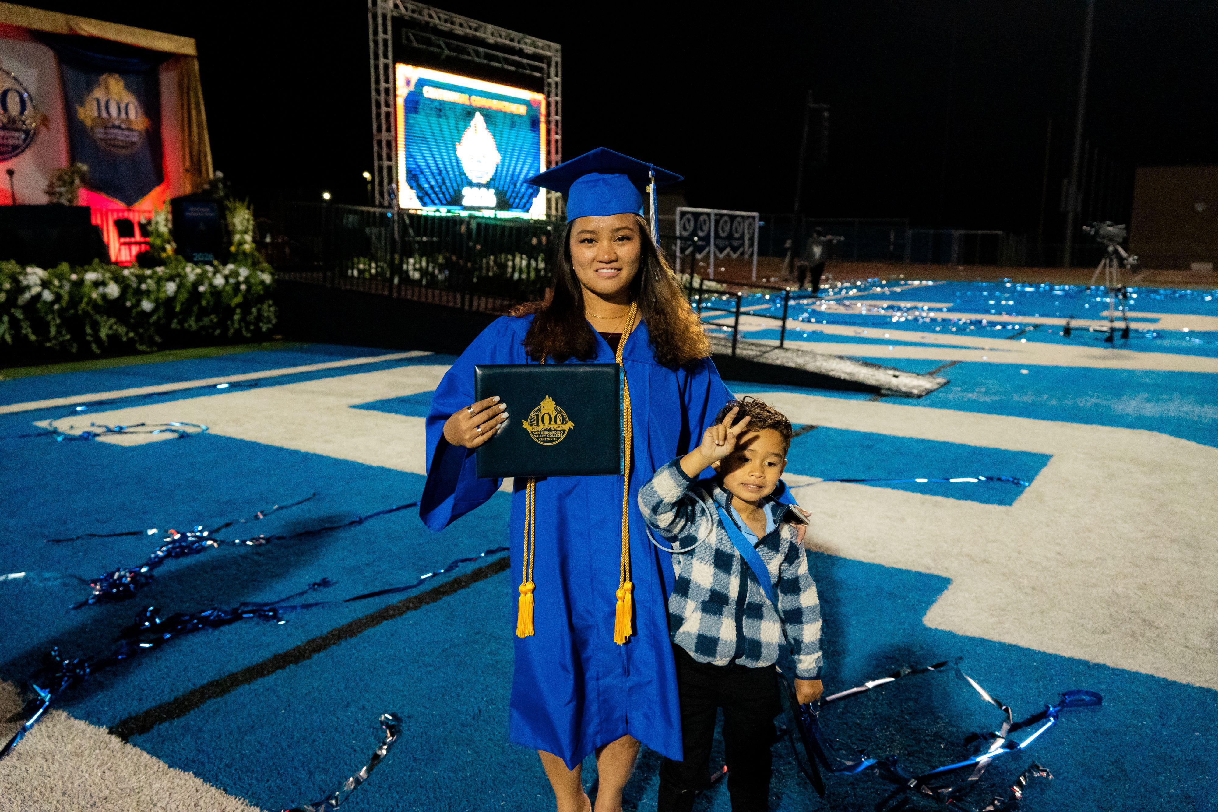 Female graduate holds diploma next to little boy in front of commencement stage