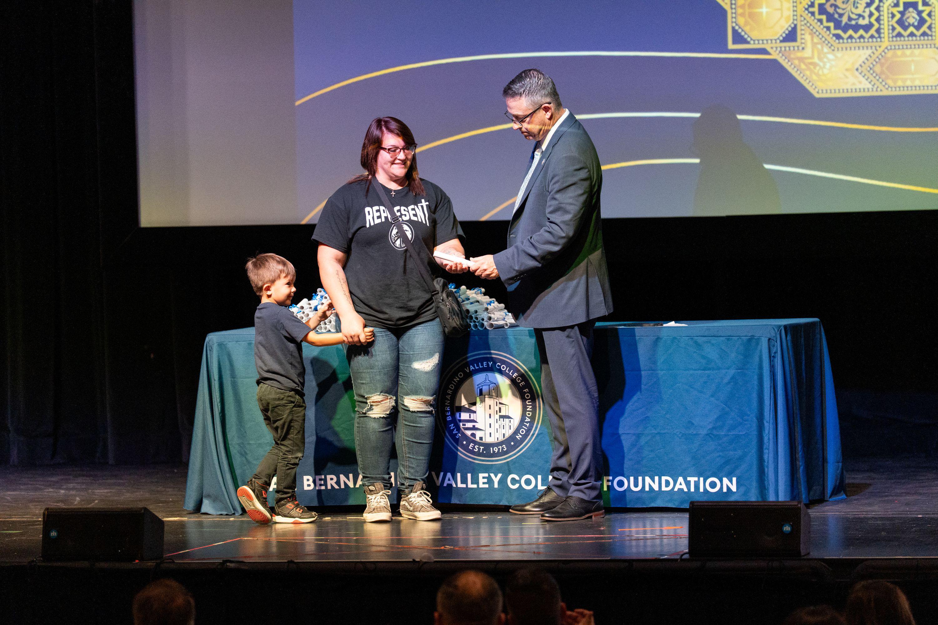 Man hands scholarship award to woman on stage as boy watches