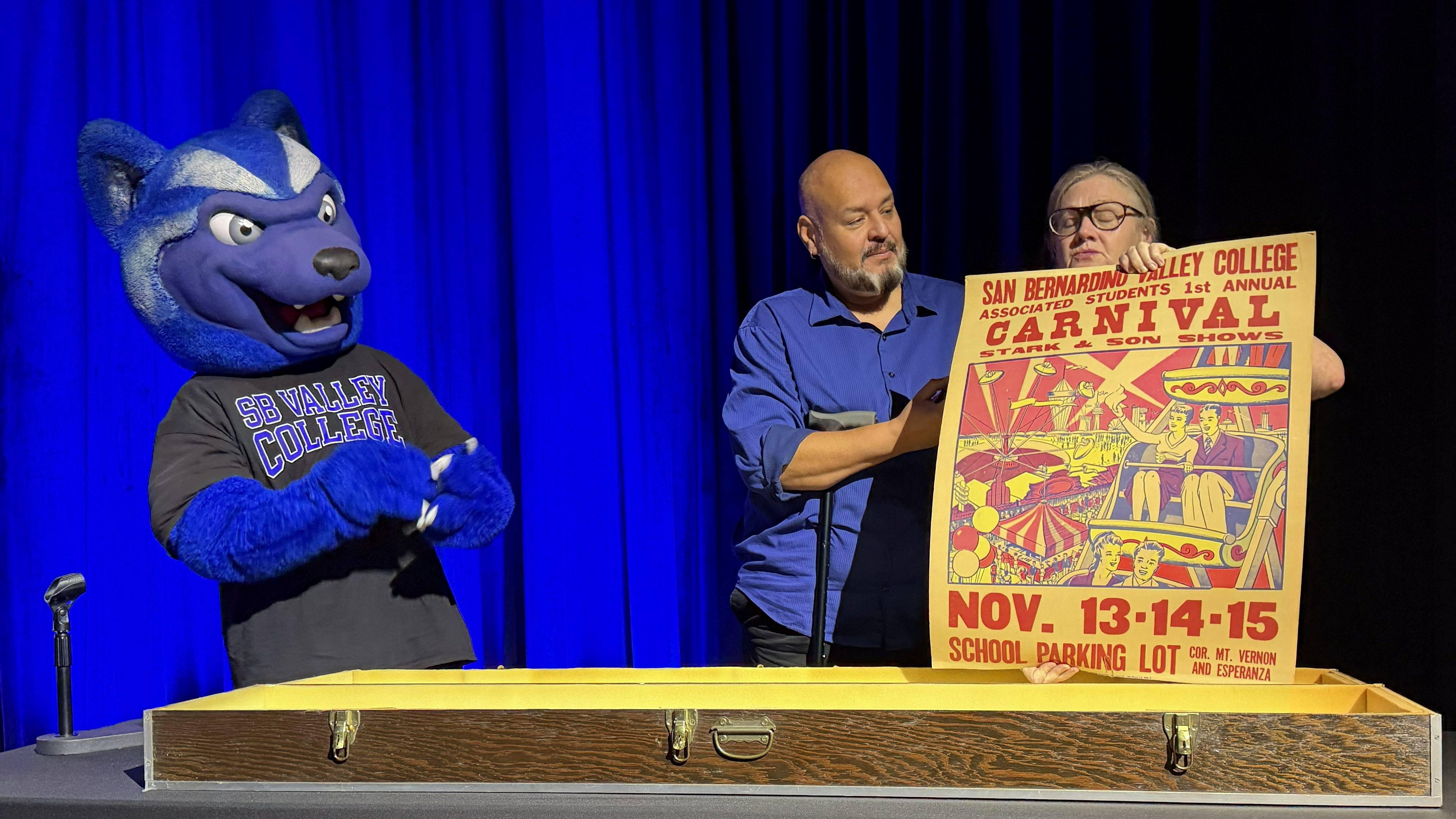 Man and woman hold 1986 poster while SBVC mascot Blue looks on