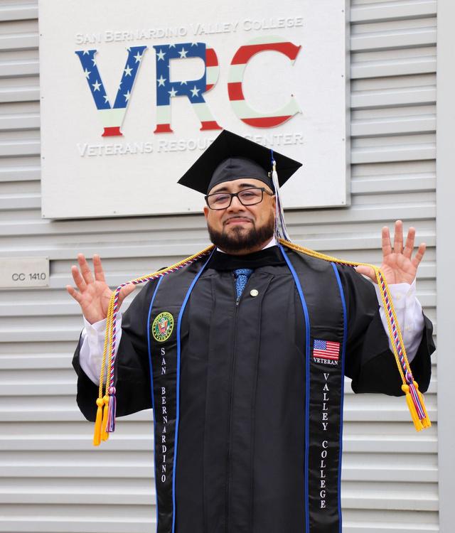 Daniel Hinojosa posing in front of the Veterans Resource Center sign in full graduation regalia.