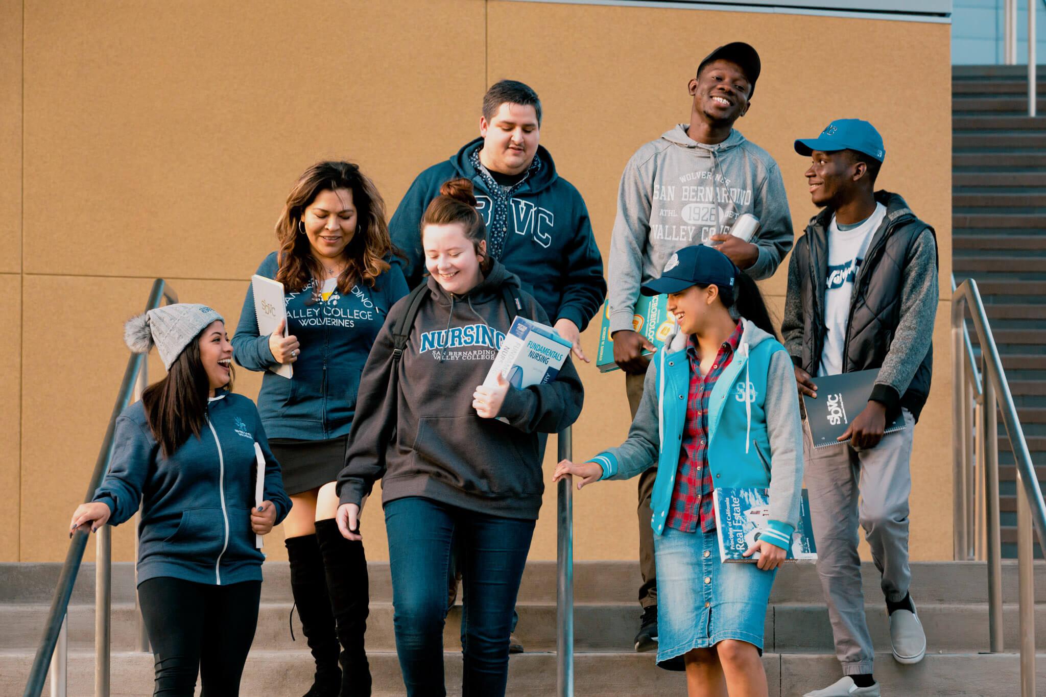 Seven SBVC students smile and walk down campus steps together, carrying textbooks and notebooks.