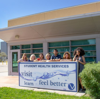 A picture of the student health services building with employees in front of the SHS sign.